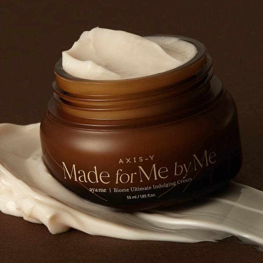 Image showing an open jar of the Axis-Y Biome Ultimate Indulging Cream. The jar is sitting on a layer of the cream. The cream is of a creamy and thick consistency and light beige in color.